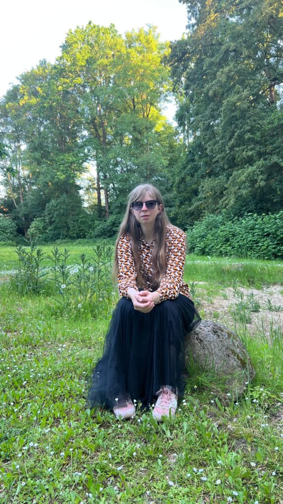 Denise Evers sitzt auf einem Stein vor Bäumen. Sie trägt eine Sonnenbrille und eine Bluse mit orange-schwarz-weißem Muster.